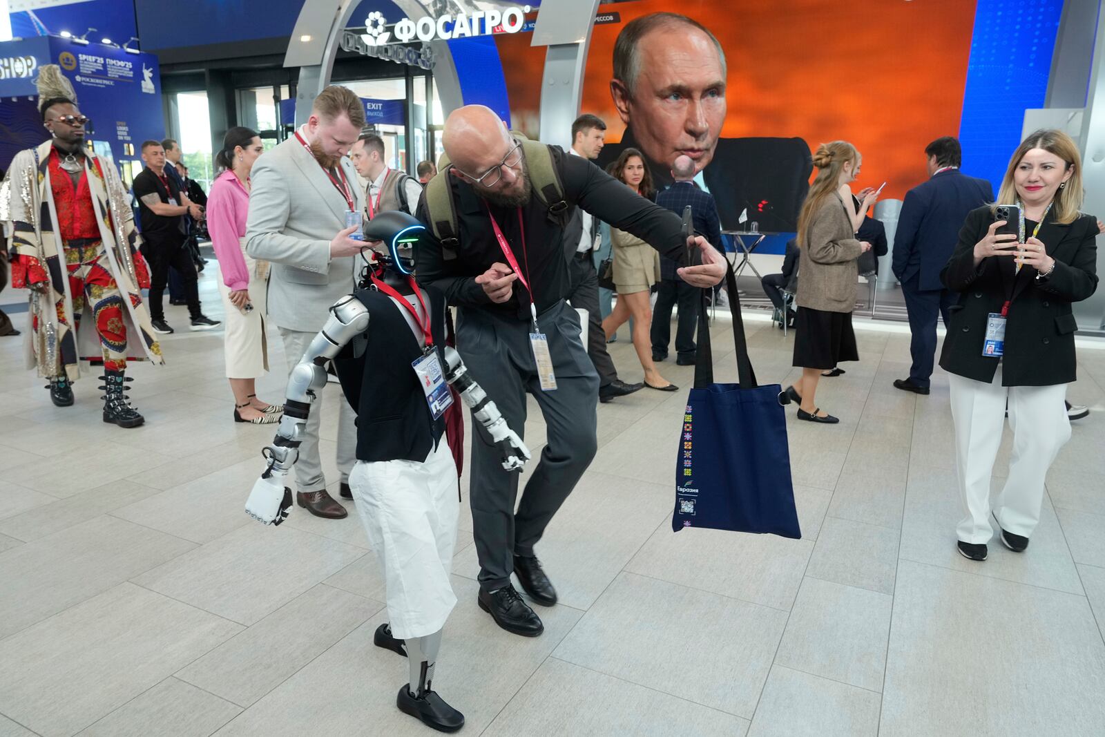 A participants makes a selfie with the Humanoid robot Volodya as other walk past a broadcast of the Russian President Vladimir Putin's speech on a TV screen during a plenary session of the St. Petersburg International Economic Forum in St. Petersburg, Russia, on Friday, June 20, 2025. (AP Photo/Dmitri Lovetsky)