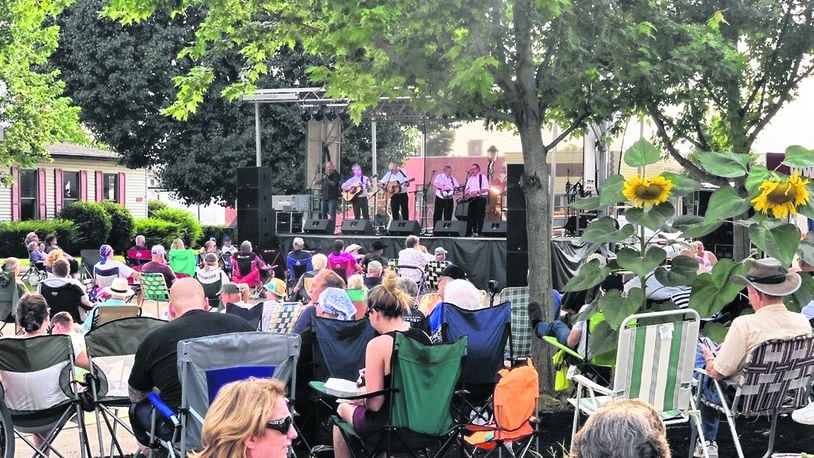A large crowd sits outside in downtown listening to a bluegrass band on stage. Some are underneath trees, and many are sitting on lawn chairs.